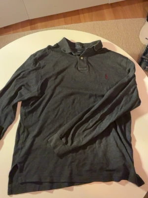 Mörkgrå pikétröja från Polo Ralph Lauren - Säljer en mörkgrå långärmad pikétröja från Polo Ralph Lauren i custom fit. Tröjan har klassisk krage, två knappar vid halsen och den ikoniska röda Polo-loggan broderad på bröstet. Perfekt till jeans och sneakers för en chill look. Modell: 175cm