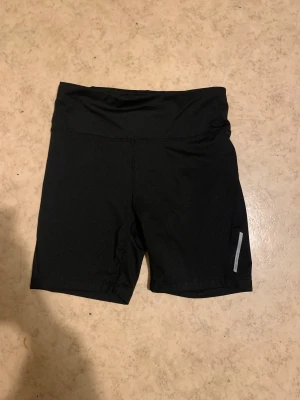 Svarta träningsshorts från SOC - Svarta träningsshorts från SOC i storlek M, tillverkade i ett tekniskt material med TCS Airflow som håller dig torr och bekväm. Shortsen har en enkel design med reflexdetalj på benet och bred linning för extra komfort.