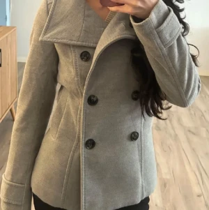 Grå kappa från H&M - Säljer en snygg grå kappa från H&M med bred krage och dubbelknäppning framtill. Jackan har långa ärmar, två sidofickor och svarta knappar som ger en clean look. Perfekt för dig som gillar klassisk stil med modern touch.