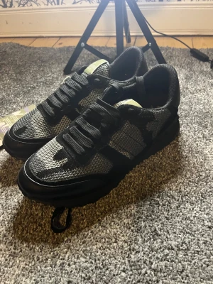 Valentino Garavani Rockrunner sneakers - Snygga Valentino Garavani Rockrunner sneakers i svart och grått med coolt camouflage-mönster. Skorna har rund tå, platt sula och snörning. Ovandelen är i mesh och skinn vilket ger en sportig vibe. Perfekta för dig som vill ha något extra till din outfit. Stl 38