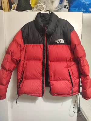 Röd och svart pufferjacka från The North Face - Säljer en fet pufferjacka från The North Face i rött och svart. Jackan har hög krage, två sidofickor med dragkedja och tydlig logga på bröstet och ryggen. Perfekt för kalla dagar med sin dunfyllda design och coola färgblock. Stängs med dragkedja framtill.