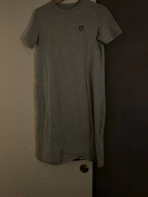 Grå t-shirtklänning från Lyle & Scott - Säljer en enkel och stilren grå t-shirtklänning från Lyle & Scott i 100% ekologisk bomull. Klänningen har korta ärmar, rund hals och en liten broderad logga på bröstet. Perfekt för en chill och avslappnad look.