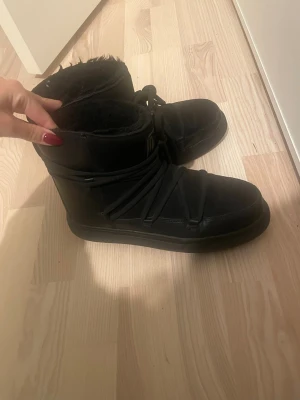 Svarta moon boots med snörning - Säljer ett par svarta moon boots med mjukt foder och snörning runt hela skon. De har platt sula och är tillverkade i syntetmaterial med pälsfoder på insidan. Perfekta för kalla vinterdagar och riktigt mysiga att ha på sig.