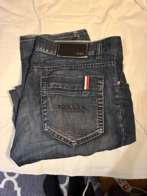 Blå jeans från Prada med broderad logga - Snygga blå jeans från Prada med broderad logga på bakfickan och coola detaljer som rödvit rand på fickorna. Klassisk femficksmodell med knapp och dragkedja framtill. Jeansen har en rak passform och är tillverkade i mjukt denimtyg. Storlek W 34 L 32