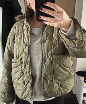Quiltad grön pufferjacka med fickor - Säljer en quiltad pufferjacka i olivgrönt med coola mönster i sömmarna. Jackan har två stora fickor framtill, tryckknappar och en hög krage. Passformen är loose och den är perfekt för dig som gillar en chill och trendig stil.