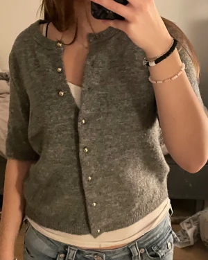 Grå stickad kofta H&M - Mysig grå stickad kofta från H&M i storlek S. Koftan har korta ärmar och snygga guldfärgade knappar framtill. Rund halsringning och ribbade kanter ger en chill vibe. Perfekt att slänga över en topp för extra lager.