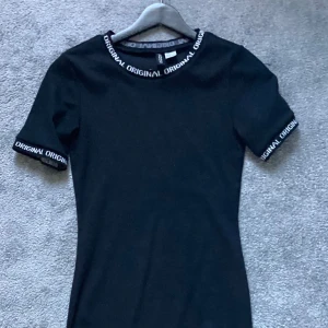 Svart klänning med textdetaljer - Snygg svart klänning med korta ärmar och textdetaljer runt hals och ärmslut. Perfekt för en stilren look.