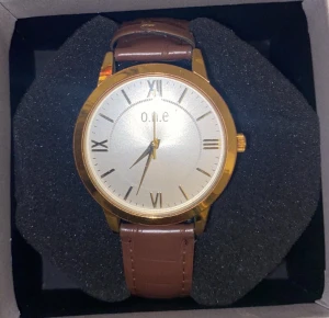 Klassisk guld klocka från o.n.e - Snygg analog klocka med rund urtavla i guld och vit bakgrund. Brunt armband i konstläder med krokodilpräglad yta och guldfärgade detaljer. Romerska siffror på urtavlan ger en stilren vibe. Perfekt för dig som gillar tidlös design.