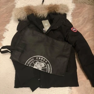 Svart Canada Goose dunjacka med päls - Säljer en svart Canada Goose dunjacka med ikonisk patch på ärmen och stor fluffig päls runt huvan. Jackan har dragkedja och knappar framtill, samt en klassisk vinterlook som håller dig varm. Perfekt för kalla dagar och riktigt snygg streetstil.