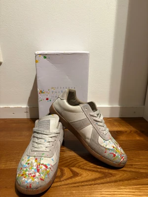 Maison Margiela sneakers med färgstänk - Snygga sneakers från Maison Margiela i vitt och beige skinn med coola färgstänk i rött, gult, grönt och blått på tån. Klassisk rund tå, platt gummisula och snörning. Boxen medföljer och har matchande färgstänk. Perfekt för dig som gillar unik design och streetstyle.