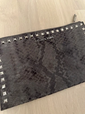 Grå kuvertväska Michael Kors med nitar - Snygg grå kuvertväska från Michael Kors med reptilmönster och silverfärgade nitar längs kanterna. Väskan har dragkedja upptill och flera fack inuti för kort och småsaker. Handtaget visar lite tecken på slitage men fungerar fint. I övrigt i mycket fint skick. Perfekt som clutch till fest eller som elegant vardagsväska. Rök- och djurfritt hem.