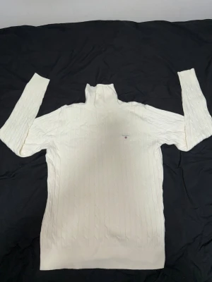 Vit polotröja från GANT i bomull - Snygg vit polotröja från GANT med kabelstickat mönster och broderad logga på bröstet. Tröjan är tillverkad i stretchig bomull och har lång ärm och ribbade muddar. Perfekt för dig som gillar klassisk och clean stil. Storlek S men den är slim