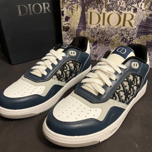 Dior sneakers i blått och vitt - Snygga Dior sneakers med marinblå och vita paneler i skinn, klassisk Dior-logga på sidan i mönstrad textil och vit platt sula. Skorna har rund tå, snörning och coola detaljer som ger en lyxig vibe. Perfekta för dig som vill sticka ut med exklusiv stil.