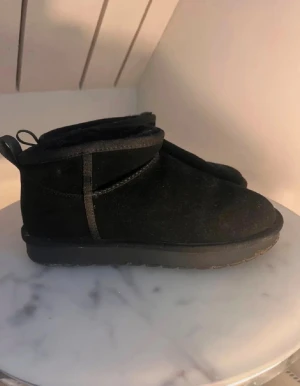 Svarta boots i mocka, storlek 40 - Svarta boots med rund tå och platt sula, tillverkade i mjuk mocka med dragflik bak. Skorna har en låg, robust siluett och grov yttersula med snöflingemönster. Perfekta för kalla dagar och har en enkel, stilren look. Minns ej vart jag köpte dom, men köpte dom för ca 700kr om jag minns rätt 