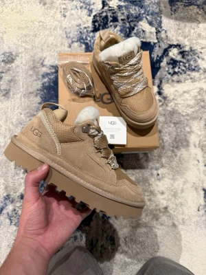 UGG Lowmel Sand  - Säljer ett par snygga beige UGG sneakers med grov platt sula och vit pälsfoder på insidan. Skorna har mockadetaljer, stickad mesh och breda snören i beige och vitt mönster. Perfekt för dig som vill ha både stil och komfort under höst och vinter.