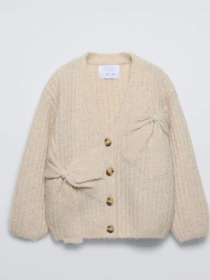 Beige kofta med rosetter från Zara - Supermysig beige kofta från Zara med stickad struktur, stora knappar framtill och två söta rosetter på ena sidan. Koftan har v-ringning och långa ärmar, perfekt för lager-på-lager under kyliga dagar.