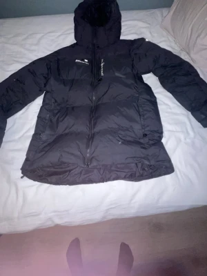 Kan snacka om pris/Svart Sail Racing dunjacka med huva - Kan snacka om pris/Svart dunjacka från Sail Racing, modell Antarctic, med huva och dragkedja framtill. Jackan har fyllning av Allied Feather + Down (500 fill power) och snygga logodetaljer på bröstet. Perfekt för kalla dagar och har en stilren, sportig look.