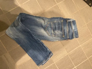 Blå Jack&Jones jeans med slitningar - Säljer ett par blå Jack&Jones jeans med snygga slitningar och ljusa tvättade partier. Jeansen har klassisk femficksdesign, smal passform och detaljer som ger en cool, använd look. Perfekta för dig som gillar en avslappnad men trendig stil!  Slim/Glenn