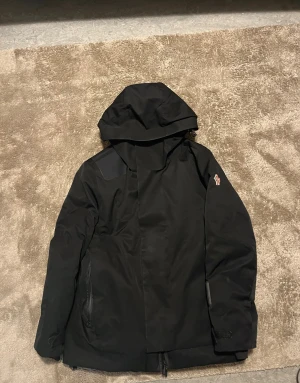 Svart dunjacka med huva från moncler - Säljer en svart dunjacka från moncler med huva och dragkedja. Jackan har en stilren look, diskret logga på ärmen och praktiska fickor med dragkedja. Perfekt för kalla dagar och har en skön, vadderad insida som håller värmen.