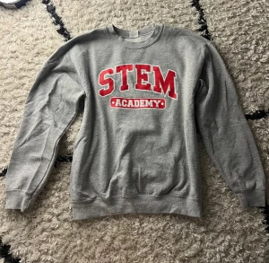 Grå sweatshirt STEM Academy - Grå sweatshirt med röd och vit text 'STEM Academy' på bröstet. Klassisk rund hals och långa ärmar, tillverkad i mjuk bomullsmix