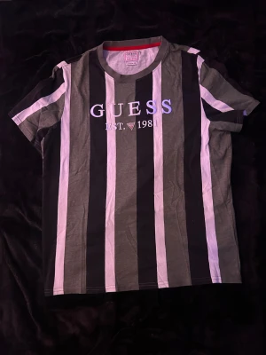 Randig t-shirt från Guess - Snygg t-shirt från Guess med breda vertikala ränder i svart, grått och vitt. Tryckt och broderad logga 'GUESS EST. 1981' på bröstet. Regular fit med rund hals och korta ärmar. Perfekt för dig som gillar streetwear och klassisk design.