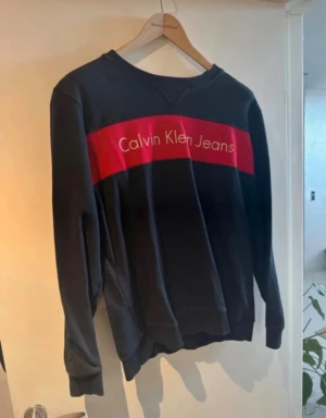 Svart sweatshirt från Calvin Klein Jeans - Svart sweatshirt med röd panel och vit logga från Calvin Klein Jeans. Tröjan har rund hals, långa ärmar och en relaxed passform. Perfekt för dig som gillar stilrena och klassiska streetwear-plagg med tydlig branding.