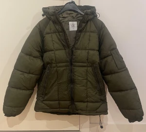 Olivgrön pufferjacka från C.P. Company - Säljer en olivgrön pufferjacka från C.P. Company med huva och dragkedja framtill. Jackan har quiltad design, två fickor med dragkedja och den klassiska C.P. Company-linsen på ärmen. Tillverkad i polyester och perfekt för kalla dagar.