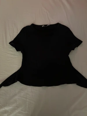 Svart ribbad topp med knyt - Säljer en svart ribbad topp med korta ärmar och rund hals. Toppen har snygga knytband i ryggen som ger en cool detalj och lite öppen rygg. Materialet är mjukt och stretchigt, perfekt för dig som gillar en slim passform och stilren look.