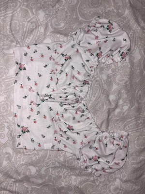 Vit blommig offshoulder topp - Säljer en söt vit offshoulder topp med små rosa och gröna blommor. Toppen har puffiga ärmar och en croppad passform med rynkade detaljer vid axlarna. Perfekt för dig som gillar romantisk och feminin stil.