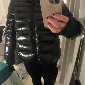 Svart pufferjacka från Colmar - Säljer en svart, glansig pufferjacka från Colmar med hög krage och dragkedja framtill. Jackan har ett stilrent, quiltat mönster och logotyp på ärmen. Perfekt för dig som vill ha en snygg och varm jacka med modern vibe.