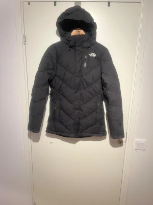 Fullrustad dunjacka från The North Face - Fullrustad dunjacka med 600-fillpower, i mycket bra skick – perfekt för kalla klimat och vinteraktiviteter. Jackan kombinerar värme, komfort och smarta funktioner för skidåkning och utomhusbruk.  Funktioner och detaljer:  • Inbyggt snölås för extra skydd mot snö    och kyla  • Avtagbar och justerbar huva  • Tjock och varm – utmärkt för kalla klimat  • Inbyggd ficka för skidglasögon (goggles pocket)  • Ficka med hål för laddare och hörlurar  • Justerbar midja för optimal passform  • Fickor på uts