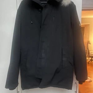 Svart dunjacka från Canada Goose black label - Svart dunjacka från Canada Goose med klassisk patch på ärmen och snygg pälsdetalj på huvan. Jackan har dragkedja och knappar framtill, två stora fickor och en stilren look. Perfekt för kalla vinterdagar och riktigt varm.* den är mer somstorlek M så stor i storleken*