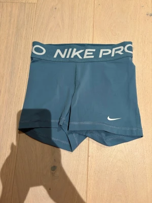 Nike Pro blå träningsshorts - Nike Pro shorts i blå färg med bred elastisk midja där det står 'NIKE PRO' i vitt. Snygg, tight passform och korta ben, perfekt för gymmet eller sport. Materialet är mjukt och stretchigt syntet som andas och sitter skönt under träning.