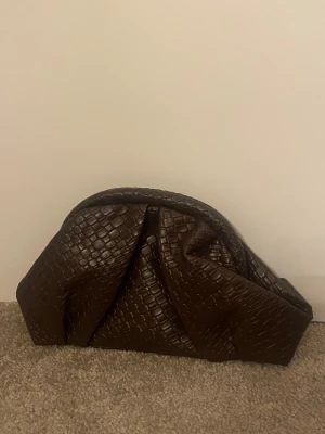 Brun kuvertväska med kedja - Helt ny!! Snygg brun kuvertväska i skinnimitation med flätad struktur och guldfärgad kedja som axelrem. Väskan har en mjuk, rundad form och stängs med magnetlås. Perfekt för att lyfta din outfit med en cool detalj.