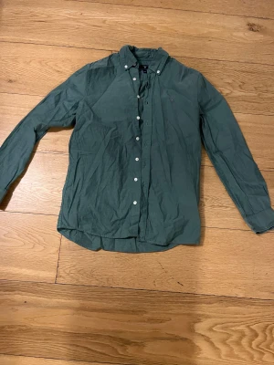 Grön skjorta från GANT med knappar - Snygg grön skjorta från GANT med klassisk button-down krage och vita knappar framtill. Skjortan har lång ärm och broderad logga på bröstet. Tillverkad i mjuk bomull som känns skön mot huden. Perfekt för dig som gillar stilrena och tidlösa plagg.