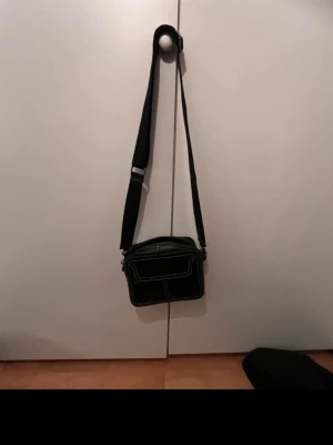 Svart axelväska med vita sömmar - Snygg svart axelväska med justerbart band och tydliga vita kontrastsömmar. Väskan har en boxig form och dragkedja upptill samt en ficka framtill. Perfekt för att bära det viktigaste och passar till många olika stilar.