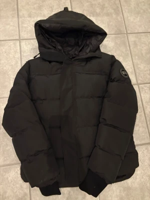 Svart pufferjacka från Canada Goose - Svart pufferjacka från Canada Goose med huva och klassisk logotyp på ärmen. Jackan har quiltad design, dragkedja och knappar framtill samt ribbade muddar vid ärmslut. Perfekt för kalla vinterdagar. (JACKAN PASSAR BÅDA L OCH M)