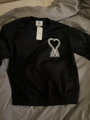 Svart sweatshirt från Ami med hjärtdetalj - Svart sweatshirt från Ami Alexandre Mattiussi med stor vit broderad hjärtdetalj och bokstaven A på bröstet. Tröjan har ribbade muddar vid hals, ärmslut och nederkant samt långärmad passform. Snyggt broderad Ami-logga i nacken.