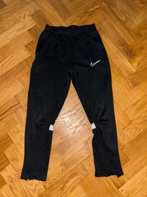 Svarta träningsbyxor från Nike - Svarta träningsbyxor från Nike med vit broderad Swoosh-logga på låret. Byxorna har resår i midjan, vita reflexdetaljer på baksidan av benen och dragkedjor vid bensluten. Tillverkade i mjukt och lätt polyestermaterial, perfekta för sport eller chill.