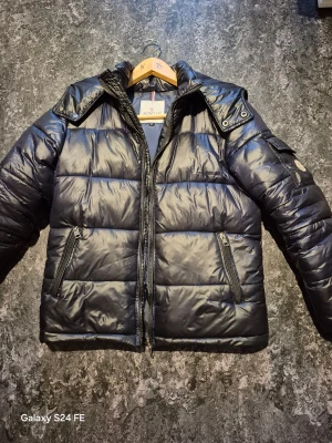 Svart Moncler pufferjacka S - Säljer en svart pufferjacka från Moncler i storlek S. Jackan har glansig finish, två dragkedjefickor fram, Moncler-logga på ärmen och quiltad design. Perfekt för kalla dagar med sin dunfyllda insida och klassiska look.
