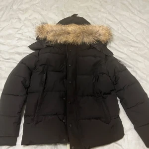 Svart dunjacka från Canada Goose - Säljer en svart dunjacka från Canada Goose med huva och snygg pälsdetalj. Jackan har broderad logga på ärmen, dragkedja och knappar framtill. Perfekt för kalla dagar med sin tjocka och quiltade design. Stilsäker och varm jacka för vintern.