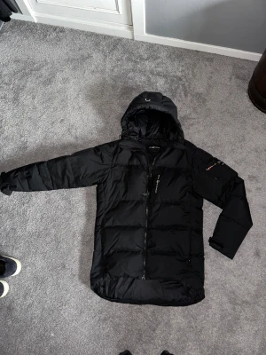 Svart pufferjacka från Sail Racing - Svart pufferjacka från Sail Racing med huva och dragkedja framtill. Jackan har en ficka med dragkedja på ärmen och flera praktiska fickor. Den är quiltad och har en clean, sportig look som funkar perfekt när det är kallt ute.  Den har en liten laglapp på vänster arm. Men syns inte!