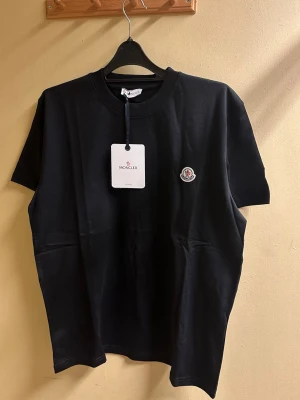 Svart Moncler t-shirt med logga - Svart t-shirt från Moncler i mjuk bomull med klassisk rund hals och korta ärmar. På bröstet sitter Monclers ikoniska logopatch i vitt och rött. Enkel och stilren design som funkar till allt. Snygg passform och exklusiv känsla.