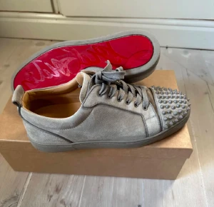 Christian Louboutin Louis Junior Spikes - Säljer ett par gråa Christian Louboutin Louis Junior Spikes sneakers i mocka med ikoniska nitar framtill och röd signatursula. Skorna har snörning och en clean, lyxig look som sticker ut. Perfekta för dig som vill ha något unikt och edgy i garderoben.
