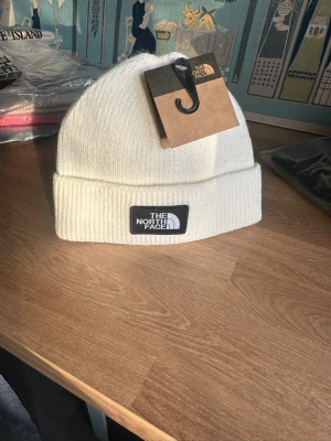 Vit ribbad mössa från The North Face - Snygg vit mössa från The North Face med ribbad struktur och uppvikt kant. Klassisk svart logopatch framtill. Perfekt för kalla dagar och enkel att matcha med din streetwear eller outdoor-look.