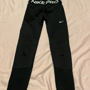 Nike Pro svarta tights - Svarta Nike Pro tights med vit logga på benet och bred resår med Nike Pro-tryck i vitt. Slimmad passform och stretchigt syntetmaterial som sitter skönt mot kroppen. Perfekta för träning eller sport.