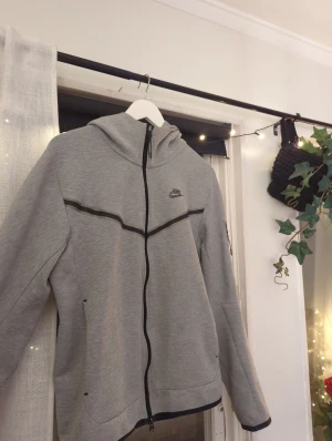 Grå Nike hoodie med dragkedja - Snygg grå hoodie från Nike med hel dragkedja och svart Nike-logga på bröstet. Hoodien har svarta detaljer längs bröstet och ärmarna samt praktisk huva. Perfekt för en sportig och avslappnad stil.