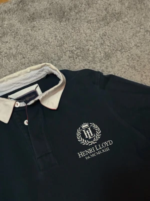Henri Lloyd pikétröja marinblå - Snygg marinblå pikétröja från Henri Lloyd i storlek S med vit krage och broderad logga på bröstet. Långa ärmar och vita knappar framtill. Stor vit siffra 6 på ryggen ger en sportig vibe. Perfekt för dig som gillar klassisk stil med en twist.