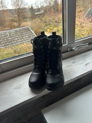 Svarta boots med klack och snörning - Säljer ett par svarta boots i skinnimitation med grov klack och snörning framtill. Skorna har rund tå, dragkedja på sidan och coola metalldetaljer runt skaftet. Perfekta för dig som vill ha en edgy stil med lite höjd.