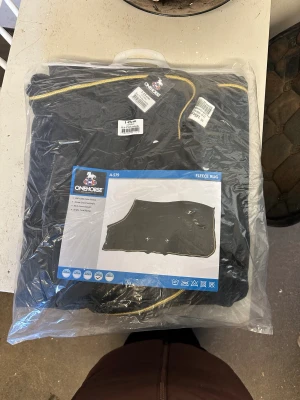 Svart fleecetäcke från OneHorse 145cm - Svart fleecetäcke från OneHorse, modell A-579, tillverkat i 250 GSM polar fleece. Täcket har guldig kant, två låga korsgjordar och frontknäppning. Perfekt för ridning och håller hästen varm under kyliga dagar. Helt oanvänt, aldrig tagits ur förpackningen. 